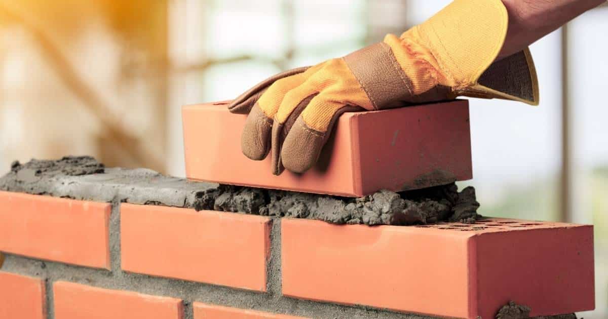 De hand van een metser in een gele leren werkhandschoen voegt een steen toe aan een gemetselde muur. De muur bestaat uit oranje snelbouwstenen en grijze metselspecie.