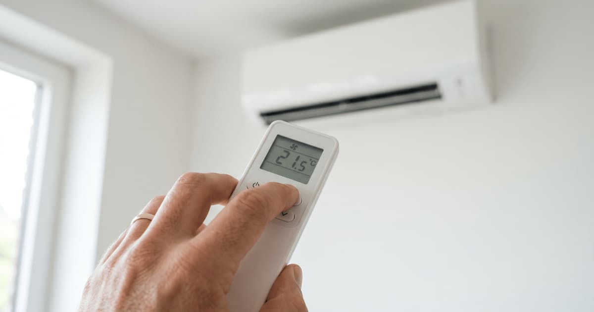 Een hand richt een witte afstandsbediening met een lcd-scherm dat 21,5 graden toont naar een wandgemonteerde airco-binnenunit in een lichte kamer met een raam op de achtergrond.