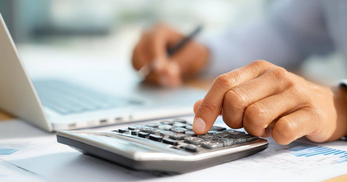 Een hand boven een rekenmachine. De wijsvinger drukt een toets in. Onder het rekenmachine ligt een document met grafieken. Op de achtergrond een laptop, met daarboven een hand met pen.