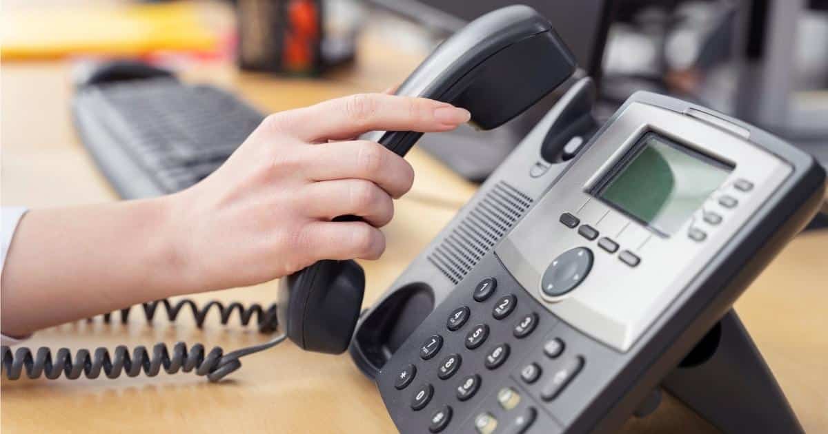 De hand van een dame neemt de hoorn van een zwart met grijs telefoontoestel met display. Het toestel staat op een bureau. Op de achtergrond zijn het toetsenbord en deel van het scherm van een computer zichtbaar.