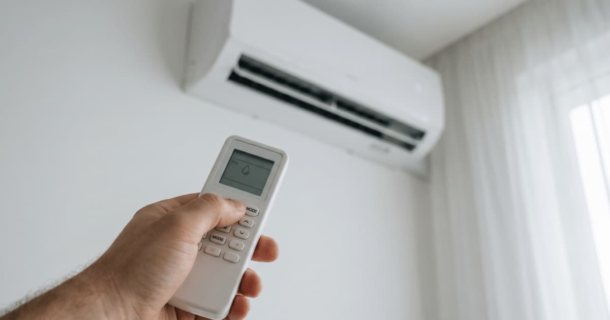 Een hand drukt op de modeknop van een witte afstandsbediening waarop een druppelsymbool op het lcd-scherm verschijnt, gericht op een airco-binnenunit aan een witte muur met een doorschijnend gordijn ernaast.