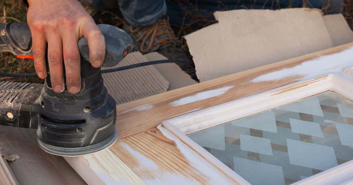 Een hand houdt een schuurmachine vast om het hout van de deur glad te maken en voor te bereiden op de schilderwerken.