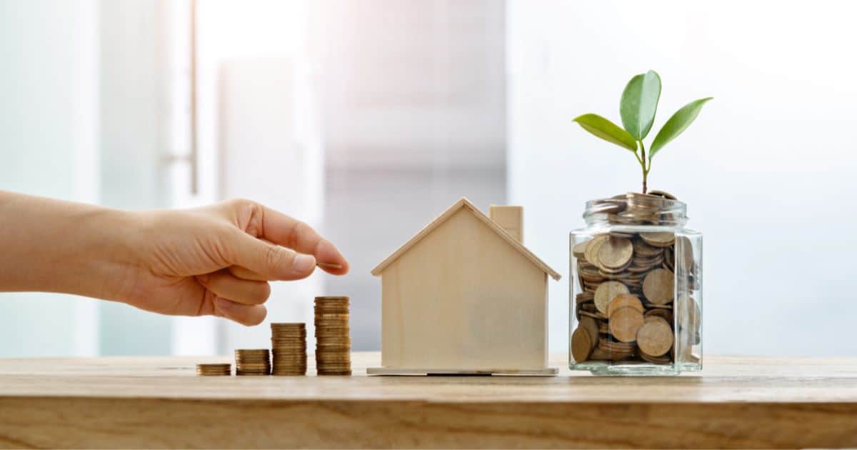 Een hand legt een muntje op een stapel centjes. Links van de stapel staan drie kleinere stapeltjes. Rechts van de stapeltjes staat een houten modelhuisje. Rechts van het huisje staat een glazen pot, gevuld met muntjes, waaruit een plantje met drie groene bladeren groeit.