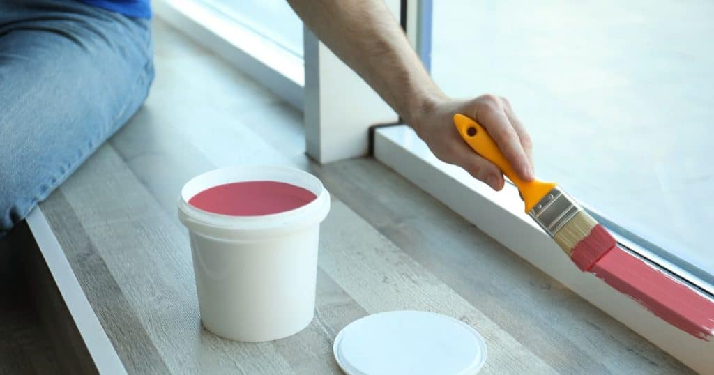 Een man of vrouw schildert met een dunne verfborstel met geel handvat de onderkant van een raamkozijn roze.