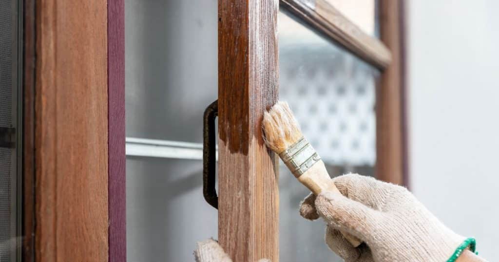 Een persoon verft of lakt een houten raam met donkere kozijnen. Hij of zij draagt daarbij beige werkhandschoenen met een fijne, groene rand.