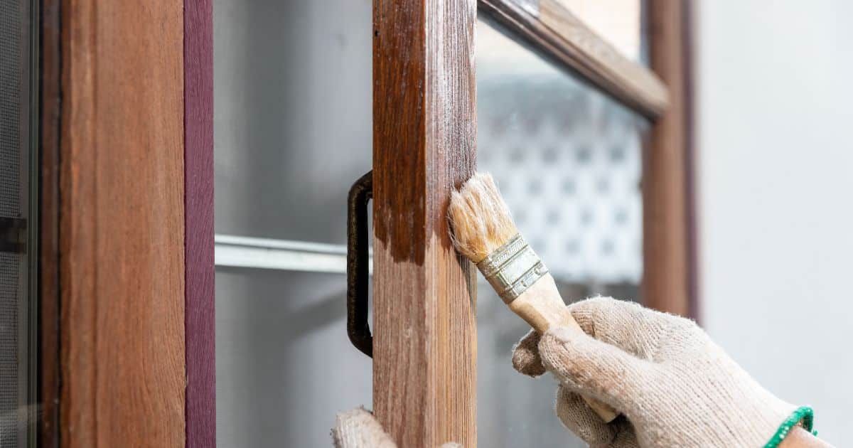 Een persoon draagt beige werkhandschoenen om een houten raam met donkere kozijnen te verven met een kleine borstel.