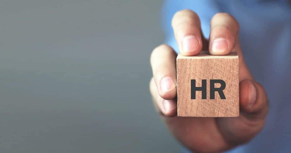 Een hand houdt een klein houten blok vast waarop de letters HR staan. Achter de hand is, vervaagd, een blauw hemd zichtbaar. De achtergrond is groengrijs.