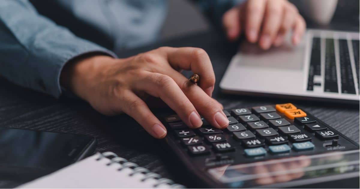 Een hand met pen boven een zwart rekenmachine. Voor de rekenmachine liggen een notitieboekje met ringen en een smartphone. Achter de rekenmachine staat een grijze laptop. Boven de mousepad zweeft een hand. De rekenmachine, de telefoon, het notitieboekje en de laptop liggen op een zwarte houten tafel.