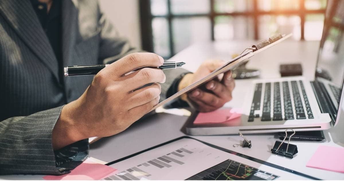 Het bovenlichaam van een man in maatpak met krijtstreep. Hij houdt een vulpen in de rechterhand. In zijn linkerhand houdt hij een klembord vast, net boven een grijze laptop. Onder het toetsenbord van de laptop kleeft een roze post-it. Naast de laptop liggen klemmetjes, en een geopende map met een document met grafieken.