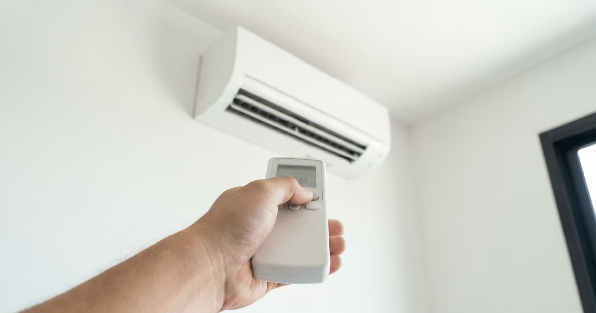 Een hand richt een witte afstandsbediening met klein lcd-scherm op een airco-binnenunit die hoog aan een witte muur hangt nabij een donker raamkozijn.