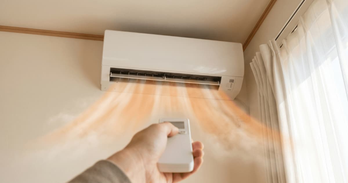 Een hand richt een witte afstandsbediening op een airco-binnenunit hoog op een crèmekleurige muur terwijl warme luchtstromen uit de geopende lamellen naar beneden blazen naast een raam met witte gordijnen.