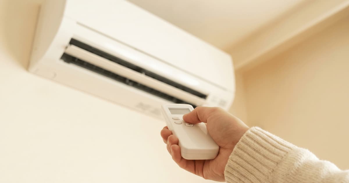Een hand in een crèmekleurige gebreide trui richt een witte afstandsbediening op een witte airco-binnenunit die hoog aan een lichtgele muur hangt.