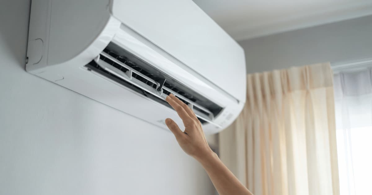 Een hand reikt omhoog naar een witte airco binnenunit met geopende lamellen en zichtbare filters tegen een lichte muur naast een raam met beige gordijnen.