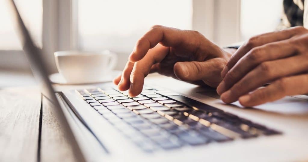 Twee handen boven het zwarte toetsenbord van een grijze laptop: ernaast staat een witte koffietas op een wit schoteltje. De tafel waarop de laptop staat, is van hout.