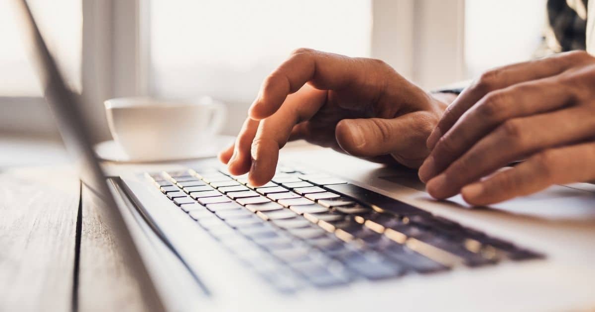 Twee handen boven het toetsenbord van een laptop. De laptop staat op een houten tafel. achter de laptop staat een wit kopje op een wit schaaltje.