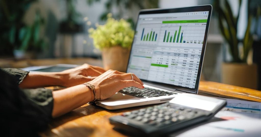 2 handen boven het toetsenbord van een zwarte laptop, met op het scherm staafdiagrammen en data in een tabel. Naast de laptop ligt een zwart rekentoestel.