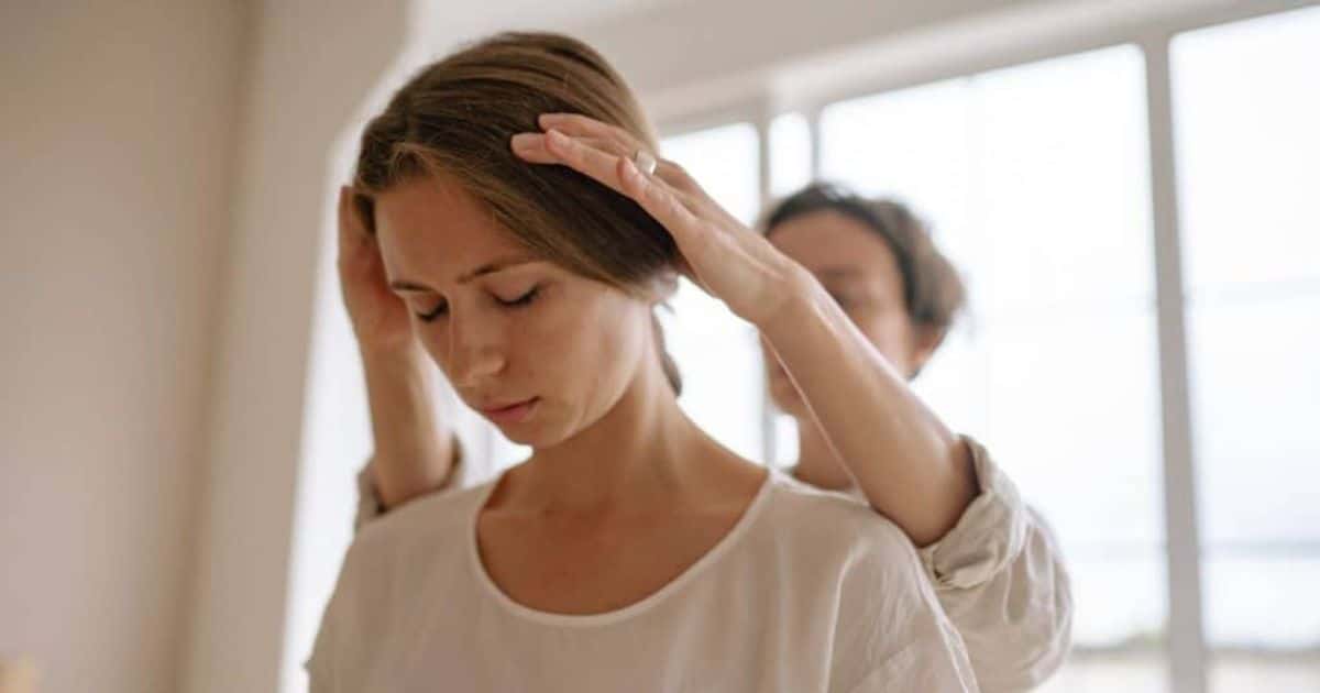 2 dames, beide dragen ze een witte blouse, staan in een soort kabinet voor een behandeling. De haptotherapeute houdt het hoofd van de patiënte vast. Die kijkt ondertussen naar beneden.