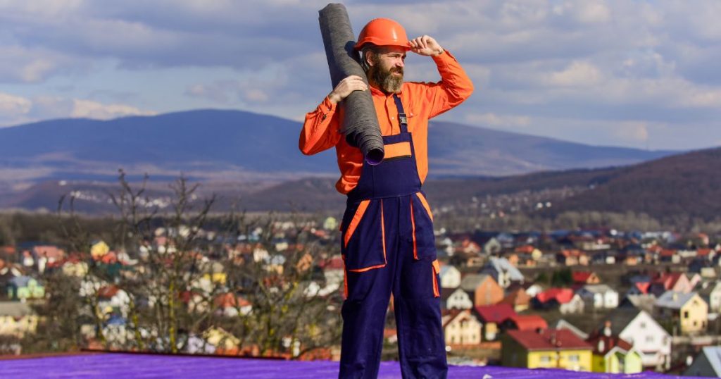 Een dakisolatiespecialist met een rol EPDM op de schouder staat zelfverzekerd op een dak. Op de achtergrond de daken van een dorp. Hij draagt een oranje hemd, oranje helm en blauwe overal.