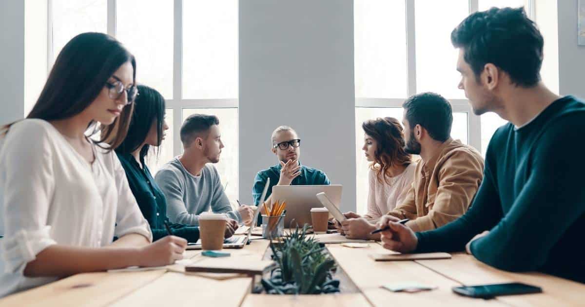 Een groep werknemers (de ondernemingsraad of afgevaardigden van de vakbond) zitten rond een lichte houten tafel. Helemaal vanachter, aan het hoofd van de tafel, zit een man met blauw hemd en zwarte bril. Links van hem zit een man met blauw hemd, naast die man zitten twee dames met lang haar. Rechts van de man aan het hoofd van de tafel zit een dame met roze trui. Naast haar zitten twee mannen met baardjes.