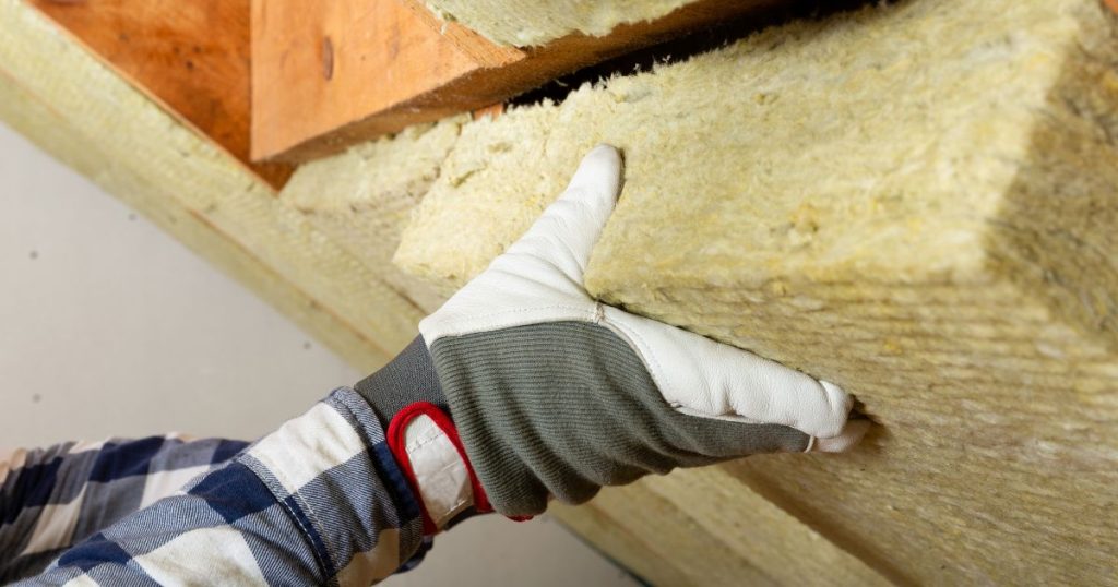 Een dikke laag zacht isolatiemateriaal tussen de kepers of gordingen van een keperdak of gordingendak. Een hand in een werkhandschoen duwt het materiaal op zijn plek.