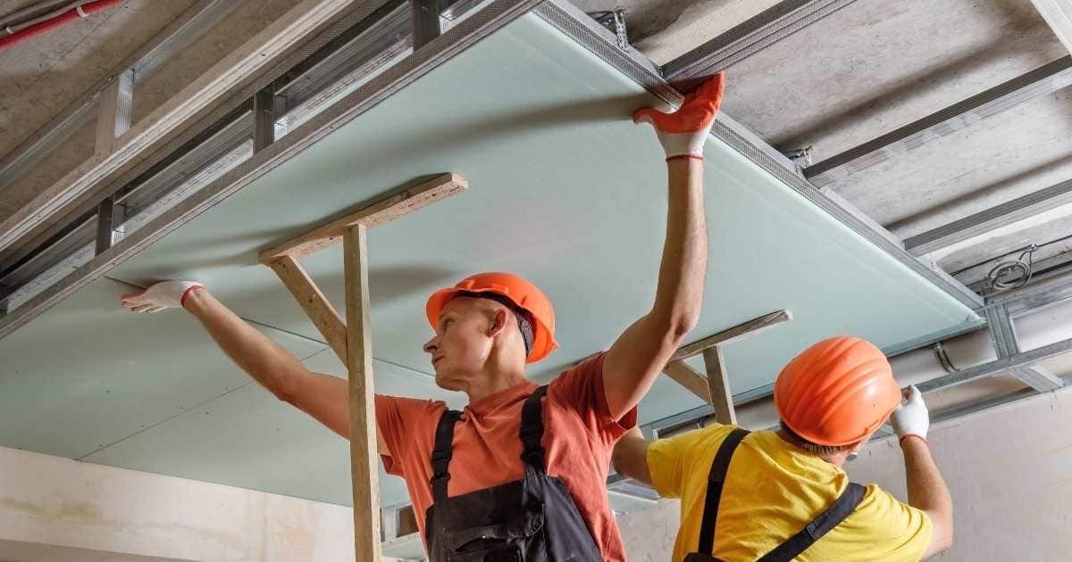 Twee gyprocspecialisten met oranje veiligheidshelmen bevestigen een gyprocplaat in een metalen draagframe onder een plafond. De voorste specialist draagt een rood t-shirt onder een zwarte overall. De andere draagt een geel t-shirt onder een zwarte overall.