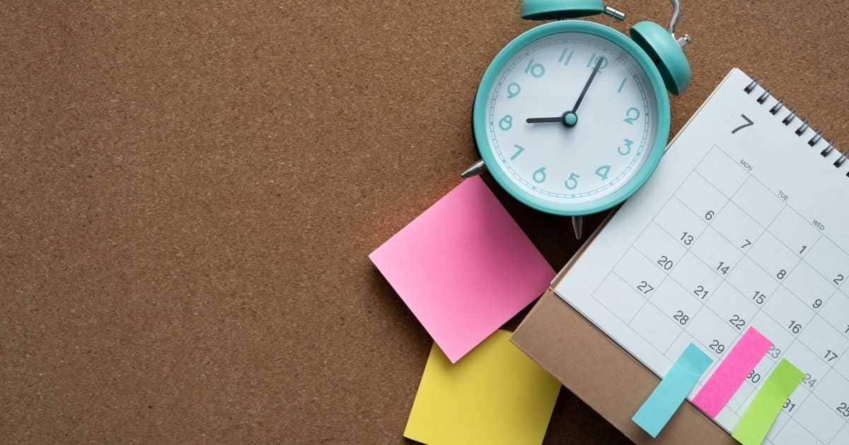 Een prikbord waarop een blauwe klok, witte agenda en stapeltjes met roze en gele post-its liggen. De wekker staat op 8 uur, de kalender (met ringen) is geopend op de 7de maand.