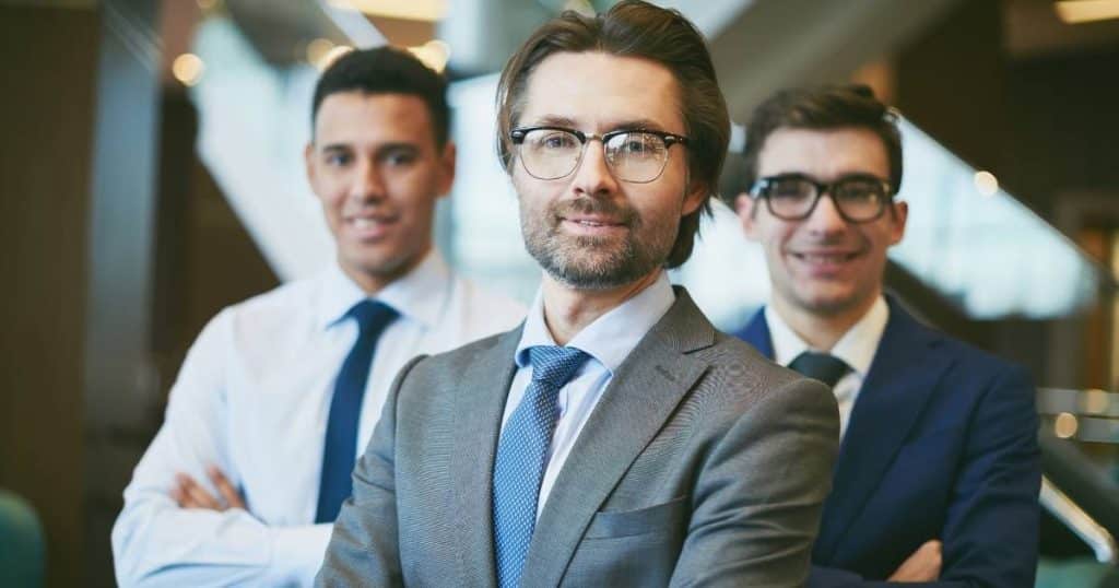 Een groep van 3 mannen met blauwe das, wit hemd en kostuumvest.