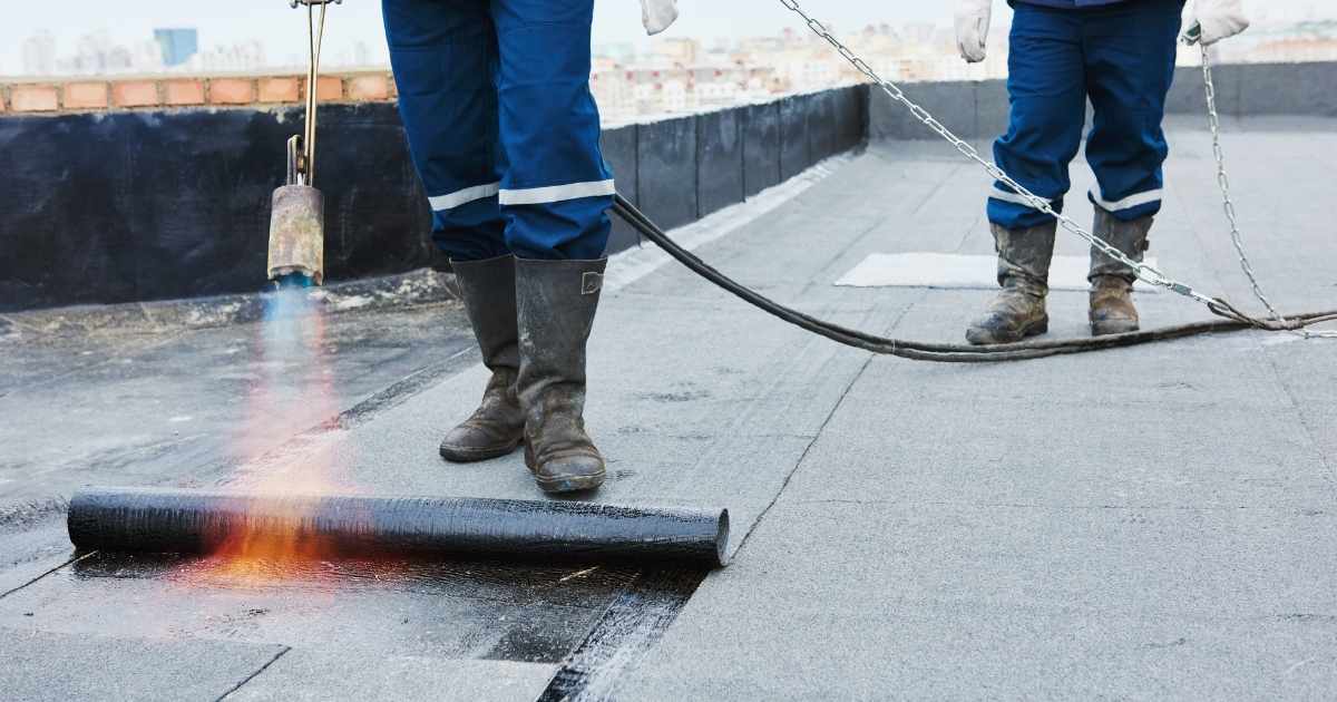 Twee dakwerkers brengen roofing aan op een plat dak met behulp van een gasbrander. De voorste dakwerker smelt de onderkant van de bitumen rol waardoor het materiaal zich goed aan de ondergrond kan hechten.