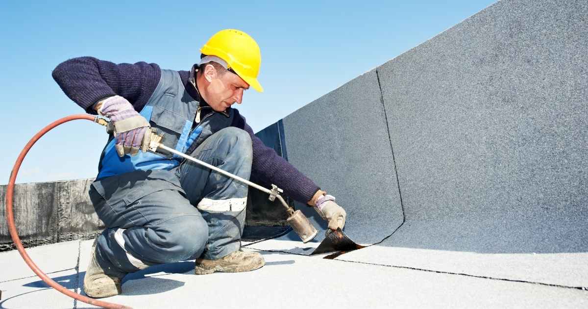 Een dakwerker met gele veiligheidshelm brengt bitumen of dakleer aan op een plat dak met een gasbrander.