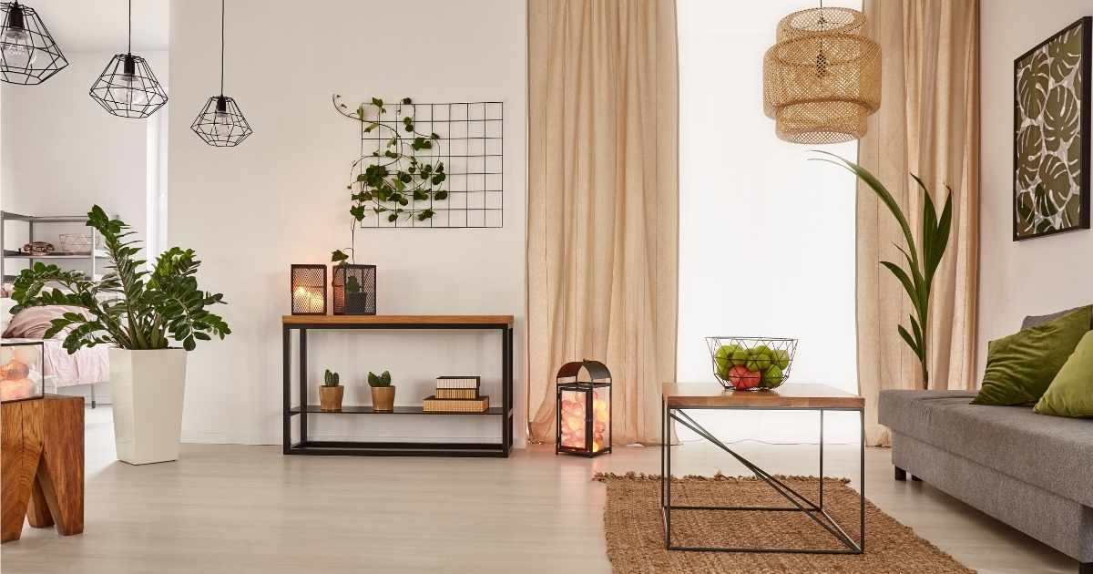 Een hippe woonkamer met houten vloer, grijze zetel met groene kussens, beige-gele gordijnen en zwarte metalen elementen. Voor de zetel staat een tafel met metalen onderstel en houten tafelblad, met daarop een metalen mand met groene en oranje vruchten. Tegen de achterste wand, naast het raam, staat een lage, zwarte metalen kast met houten blad. In de kast staan potplantjes en ligt een stapeltje met 3 boeken. Er staan verschillende grotere kamerplanten in de ruimte.