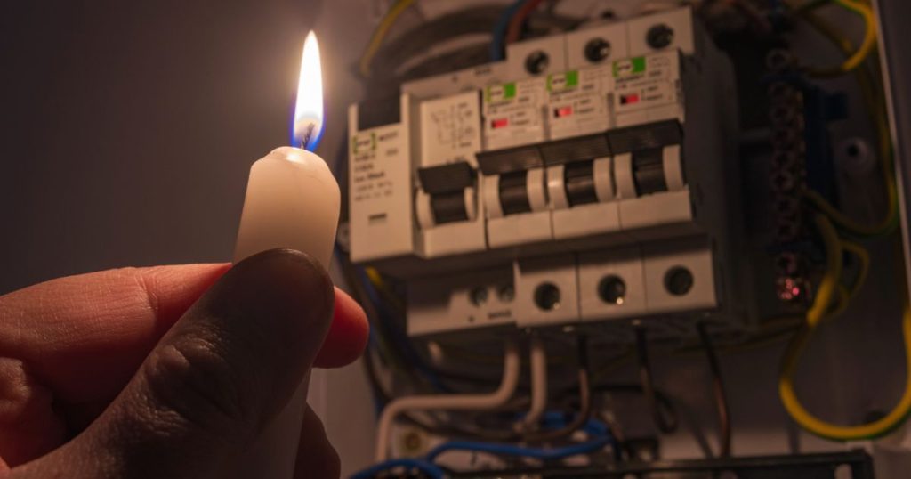 Een hand houdt een witte brandende kaars vast met zijn vingertoppen. De kaars belicht de elektriciteitskast. Rond de elektriciteitskast hangen verschillende draden.