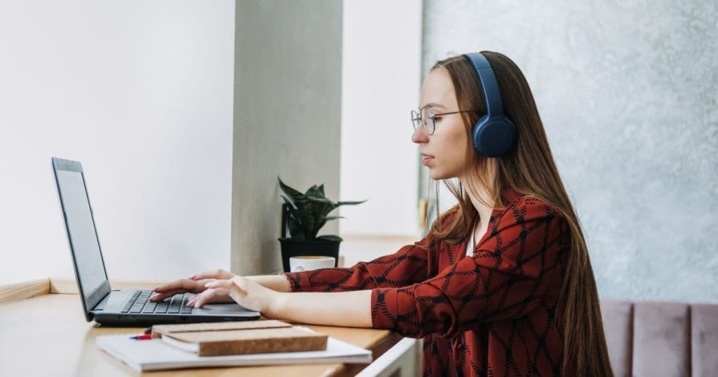 Vrouw met rode trui aan een bureau met laptop en notitieboekjes. Ze draagt ook een blauwe hoofdtelefoon.