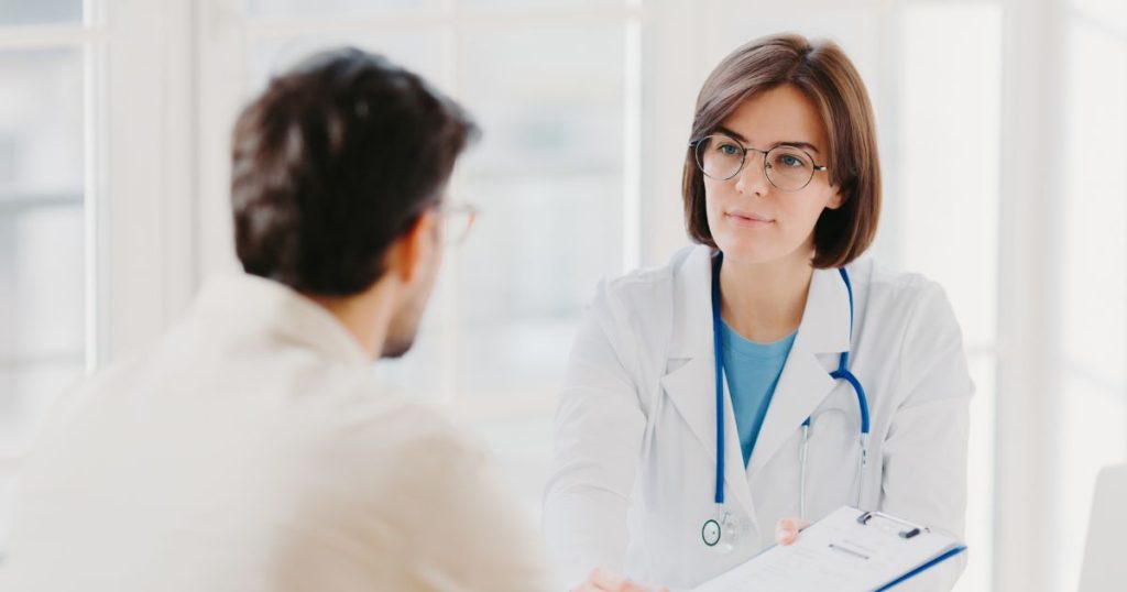 Een dokter met bril en kort bruin haar in een witte doktersjas met stethoscoop rond de schouders en een blauw t-shirt houdt een klembord voor aan haar mannelijke patiënt met wit hemd en bril.