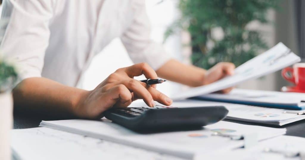 Een persoon in wit hemd typt met zijn rechterhand op een rekentoestel. In de linkerhand houdt hij een wit blad vast. Op de tafel staat ook een rode koffietas.