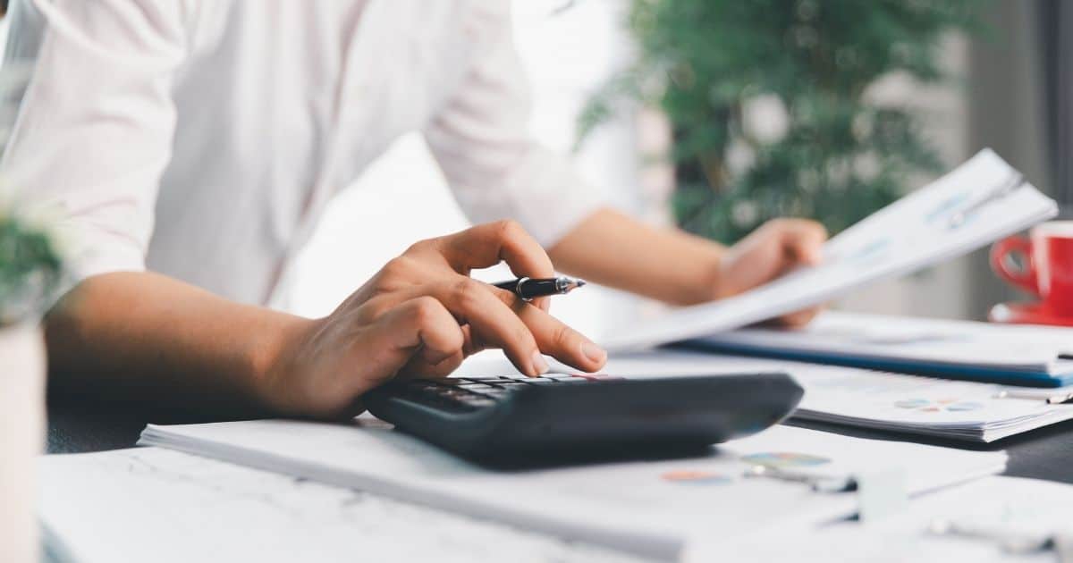 Een dame laat haar hand boven een rekenmachine hangen. In de andere hand heeft ze papieren vast. Op de tafel onder haar liggen nog meer papieren. De dame draagt een wit hemd: enkel haar armen en bovenlichaam zijn zichtbaar.