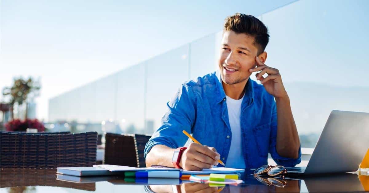 Een man met kort bruin haar, een stoppelbaard en een wit t-shirt onder een blauw hemd kijkt naar zijn rechterkant. Hij houdt zijn linkerhand tegen zijn gezicht. In zijn rechterhand houdt hij een potlood boven een notitieblok dat naast zijn laptop ligt. Voor hem op tafel liggen een bril, een stapel post-its, stiften en papieren. Hij zit buiten op een terras met een glazen scheidingswand.