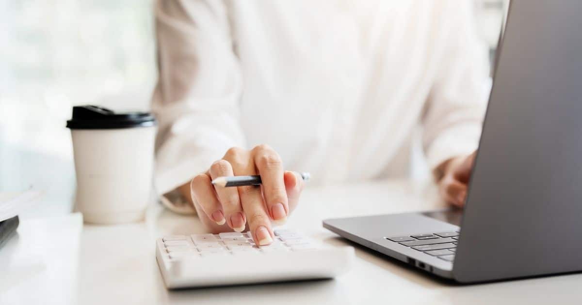 Een dame in witte trui en Franse nagels houdt haar rechterhand boven een wit rekenmachine. Tussen haar duim, wijsvinger en middelvinger houdt ze een zwart potlood. Haar linkerhand zweeft boven een grijze laptop. Naast haar rechterelleboog staat een witte wegwerpbeker met zwart deksel.