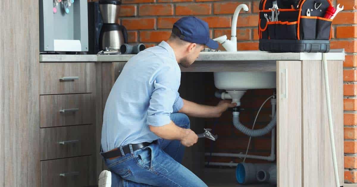 Een loodgieter met een lichtblauw hemd, blauwe jeansbroek en donkerblauw petje werkt aan de afvoer van een gootsteen in een keuken. De witte gootsteen zit in een natuurstenen aanrecht, op een houten aanrechtblok. Achter het aanrecht staat een oranje bakstenen muur.