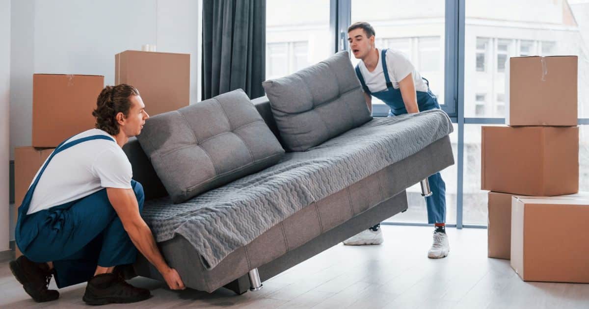 Twee professionele verhuizers verplaatsen een grijze zetel in een ruimte met grijze vloer. Ze dragen witte t-shirts en blauwe overalls. Naast hen staan stapels met kartonnen verhuisdozen.