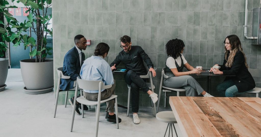 Een groep van 5 jonge collega's zit in de cafetaria van het werk tijdens de lunch. Ze zitten in 2 groepjes aan zwarte tafeltjes op grijze stoelen, tegen een wand met grijze, verticale tegels. In de linkerhoek staan 2 potten met grote kamerplanten.