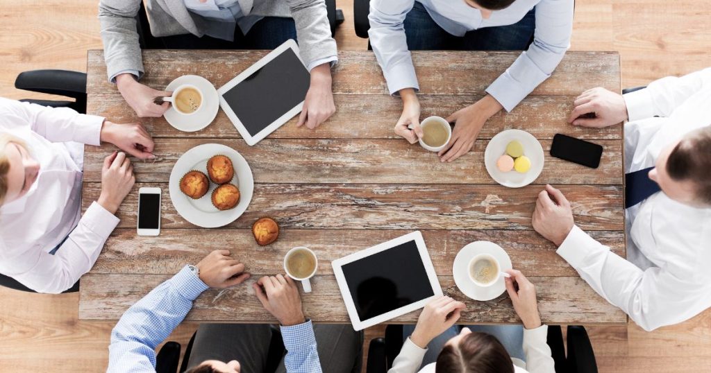 Bovenaanzicht van 6 collega's rond een houten tafel die samen genieten van koffie en gebakjes. Op tafel liggen 2 tablets, 2 smartphones, een bord met cupcakes, een bord met macarons, 4 koppen koffie, 2 koffiebordjes, en de handen van alle collega's.