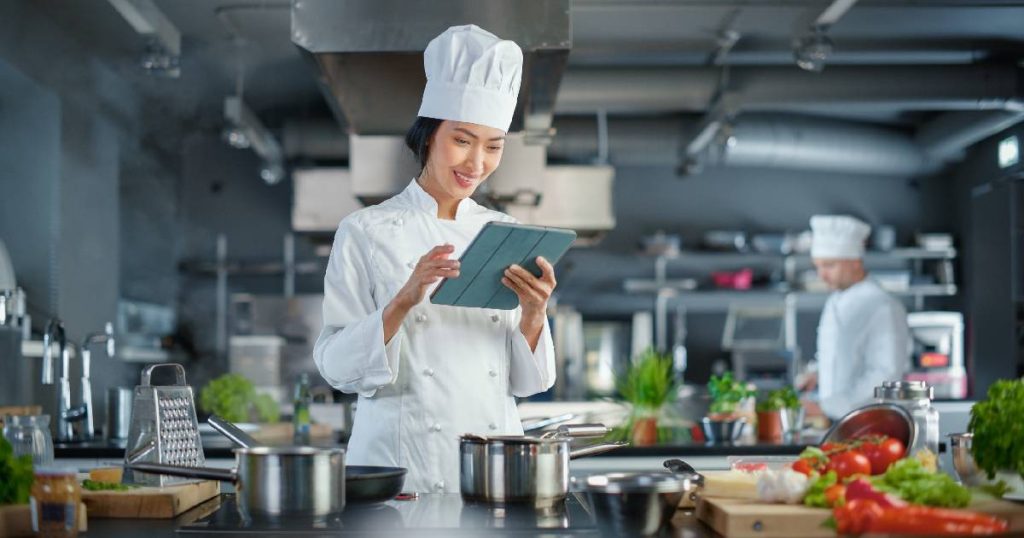 Een vrouwelijke chef gebruikt een hand-held extensie van een horeca kassasysteem om een bestelling te controleren. De chef bevindt zich in een ruime restaurantkeuken.