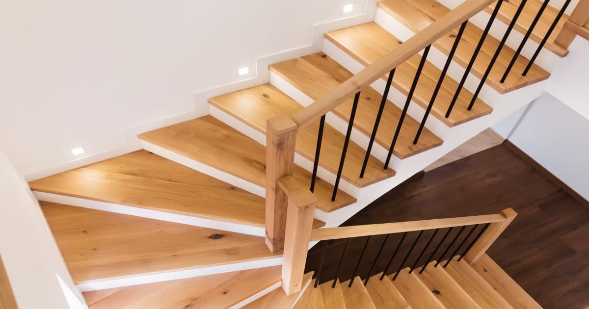 Een houten trappenhal met een houten trapleuning en zwarte accenten.  De vloer op de benedenverdieping is van een ander type hout dan de traptreden.