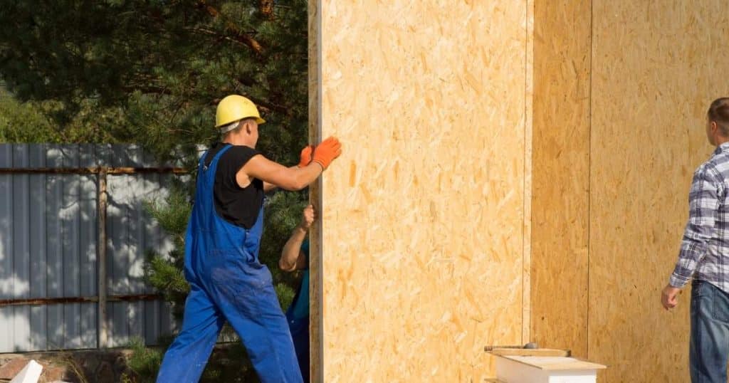 Twee vakmannen zijn in de weer met het plaatsen van de houten wanden van een prefab aanbouw. De eigenaar van de woning kijkt toe.