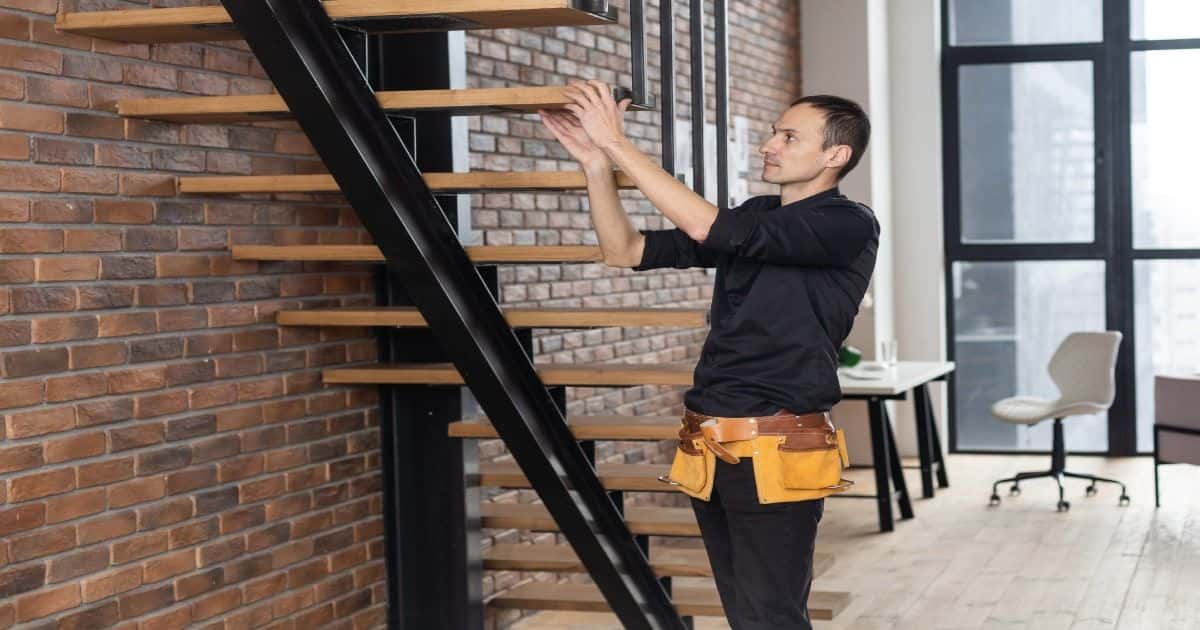 Een trappenbouwer controleert de houten traptredes die bevestigd zijn aan een metalen of stalen balk. Net als de trap zelf oogt het interieur op de achtergrond vrij industrieel.