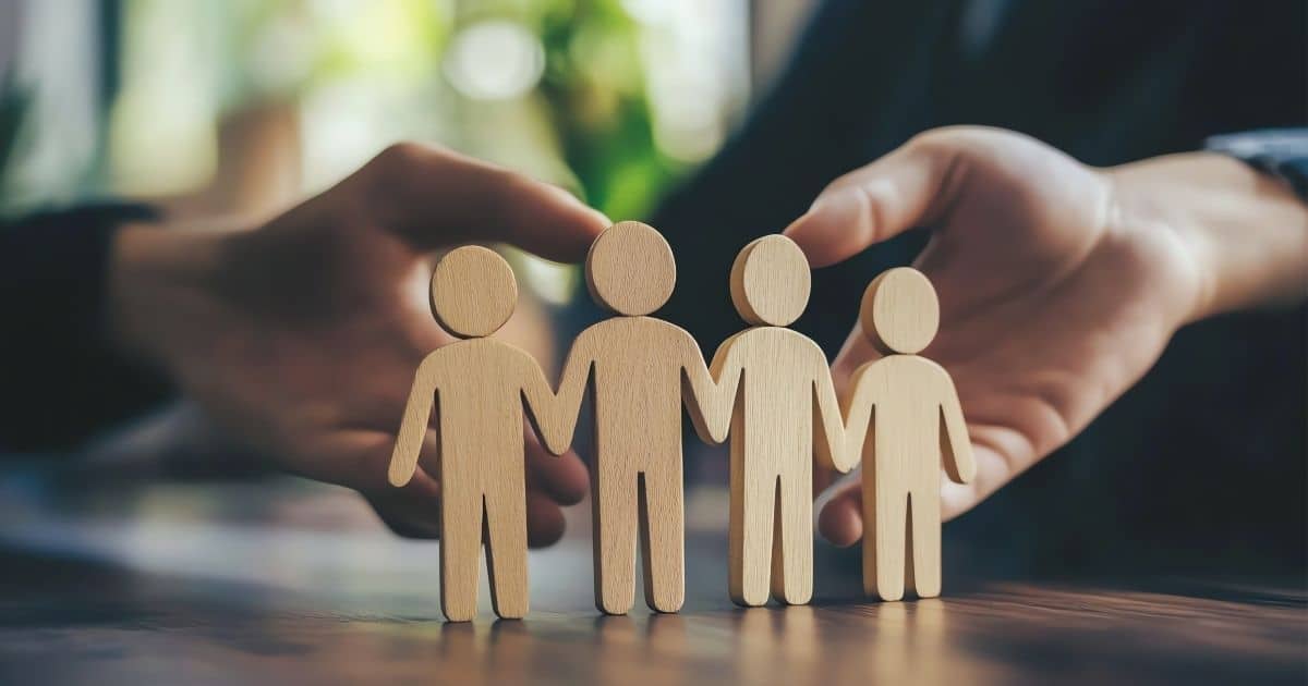 Vier uit hout gesneden menselijke figuurtjes op een rijtje met de handen tegen elkaar. Twee handen houden de poppetjes rechtop. De poppetjes staan op een houten tafelblad.