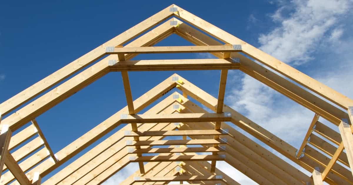 Een houten dakconstructie van een gordingendak in aanbouw toont parallelle kepers die naar de nok lopen met horizontale gordingen en metalen verbindingsplaten op de knooppunten tegen een helderblauw lucht met lichte wolken.