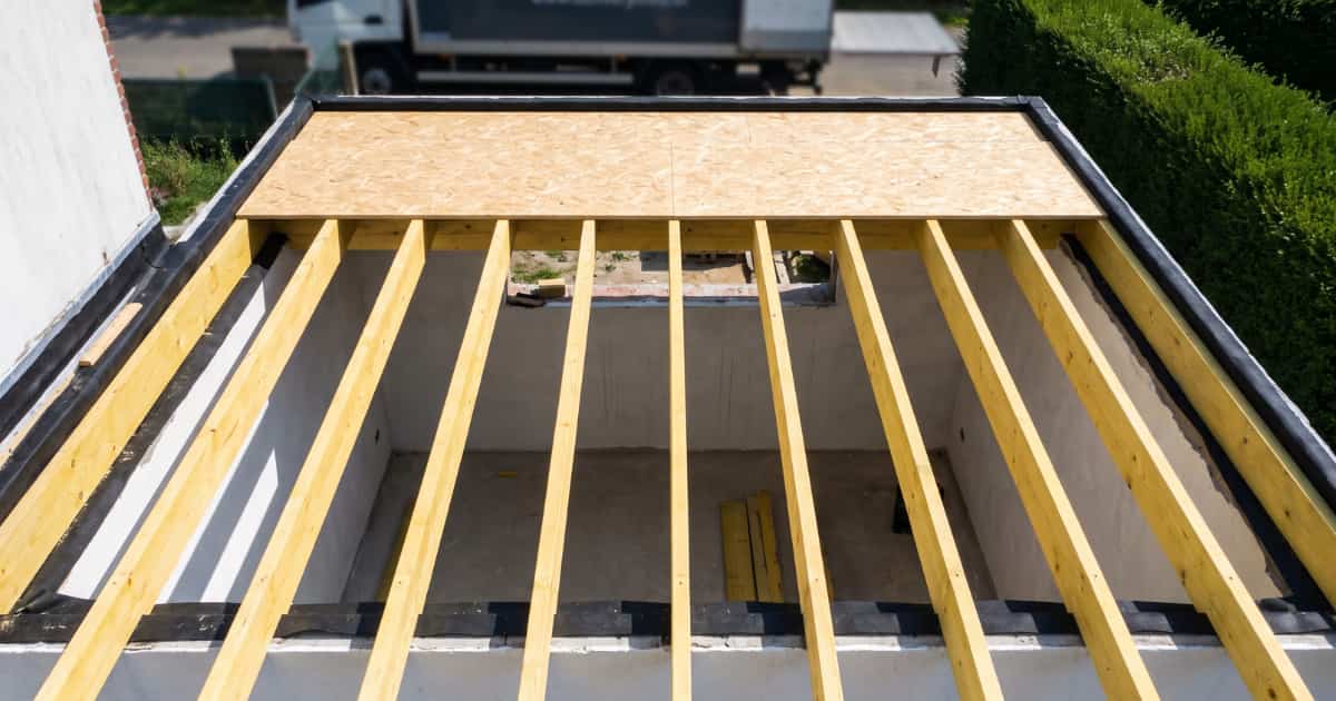 Bovenaanzicht van een plat dak in opbouw met evenwijdig geplaatste houten balken op bepleisterde muren waarbij een OSB plaat gedeeltelijk de roostering bedekt en zwarte dakfolie zichtbaar is langs de randen.