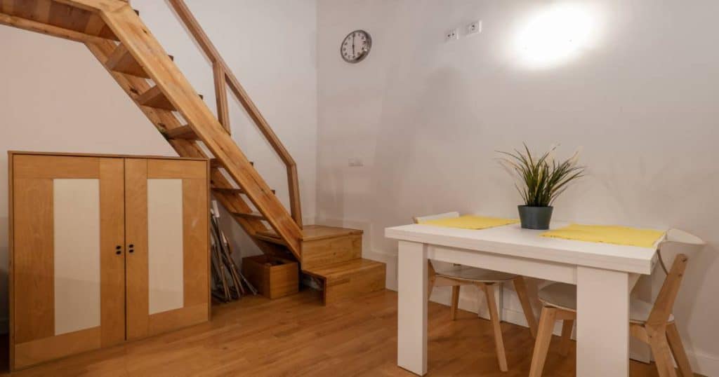 Een leefruimte met centraal een witte tafel met brede tafelpoten: tegen de muur staat een houten trap. Onderaan de trap staat nog een bordes die de overgang maakt naar de leefruimte.