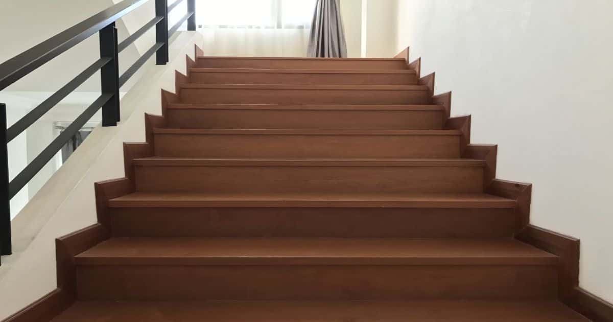 Een brede trap in donker hout: de treden en de plinten zijn in dezelfde donkere kleur. De zwarte trapleuning is van metaal.